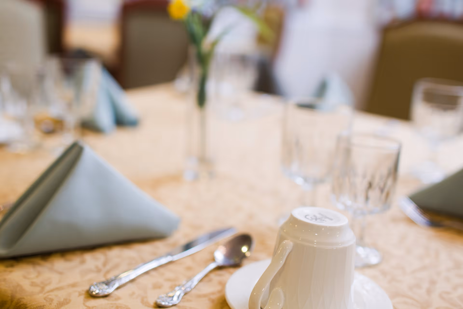 Close-up view of a dining table set with folded light blue napkins, clear drinking glasses, silverware, and an upside-down white coffee cup on a saucer. A vase with yellow flowers is visible in the background.
