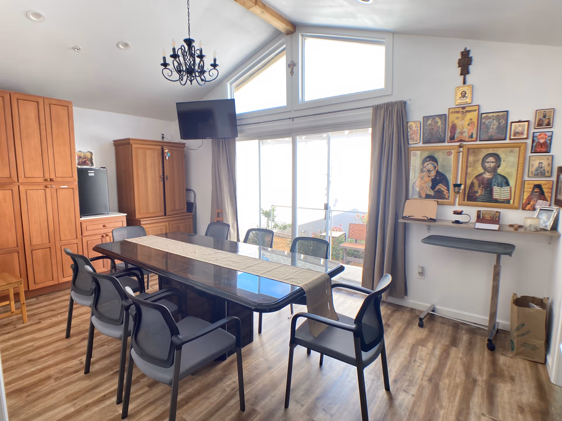 A bright dining room with a long rectangular table surrounded by eight chairs. The room features wooden cabinets, a small refrigerator, a wall-mounted TV, and a chandelier hanging from the ceiling. Large windows with curtains allow natural light to enter, and a wall displays several religious icons and crosses. The floor is wooden, and there is a small rolling table and a paper bag in the corner.