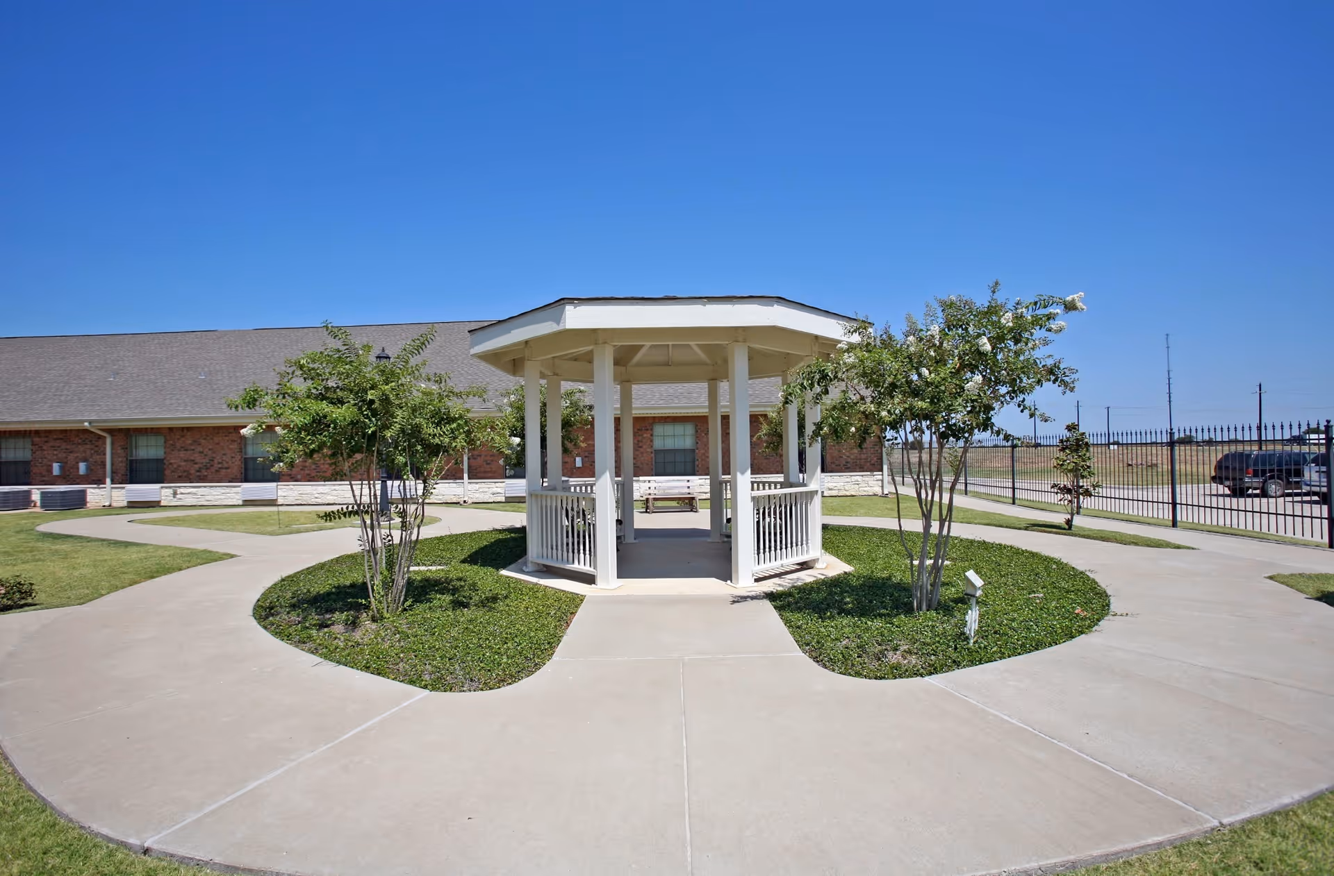 Outdoor area of Royse City Medical Lodge featuring a white gazebo surrounded by a circular concrete walkway and green grass with small trees, under a clear blue sky.