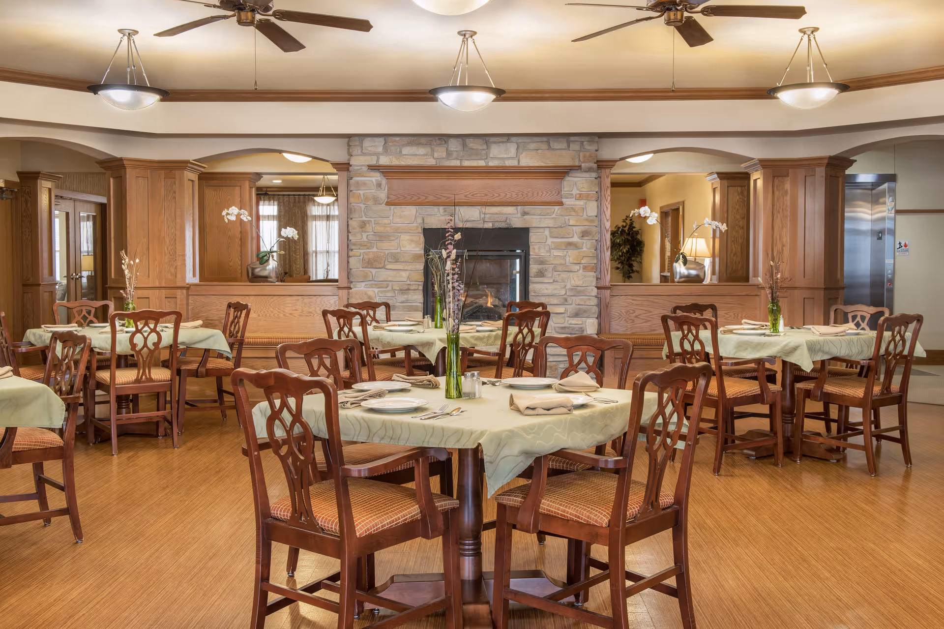 A dining room with several round tables covered with light green tablecloths, each set with plates, napkins, and silverware. Wooden chairs with cushioned seats surround the tables. The room features a stone fireplace in the center background, wooden paneling, ceiling fans, and hanging light fixtures. There is an elevator visible on the right side of the room.