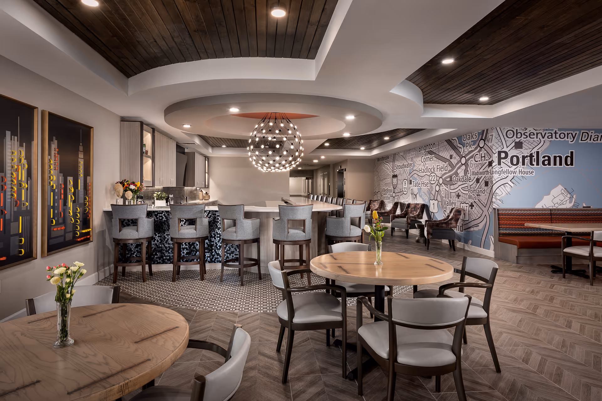 A modern dining and lounge area featuring round wooden tables with chairs, a bar with high stools, and a decorative spherical chandelier hanging from a ceiling with wooden paneling. The back wall displays a large map of Portland with various neighborhoods and landmarks labeled. There are also framed artworks on one wall and a seating area with armchairs and a booth along the wall.