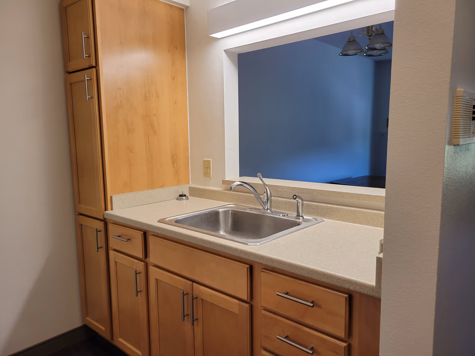 A kitchen sink area with a stainless steel sink and chrome faucet set in a beige countertop. Light wood cabinets and drawers are below and beside the countertop. A large pass-through window opens to an adjacent room with blue walls and a ceiling light fixture.