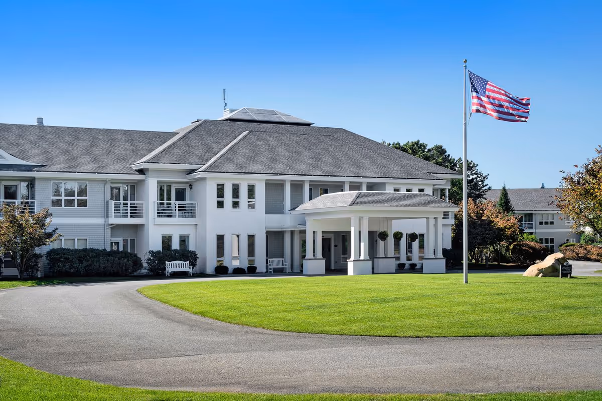 Exterior view of a two-story senior living facility building with a covered entrance, well-maintained green lawn, an American flag on a flagpole, and clear blue sky.