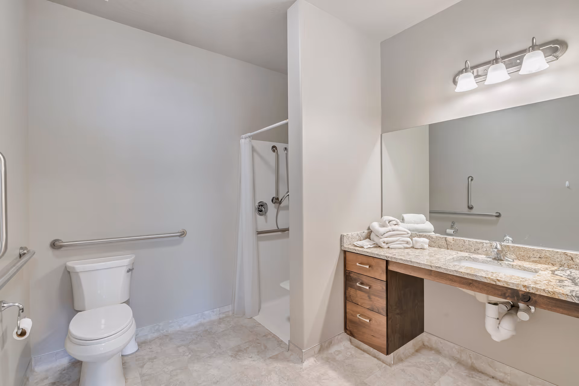 A clean, accessible bathroom with a white toilet, grab bars on the walls, a walk-in shower with a white curtain and grab bars, and a vanity with a granite countertop, sink, and a large mirror above. Several folded white towels are placed on the countertop.