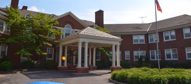 Brick senior living facility front entrance with a covered portico, white columns, landscaping, and an American flag.