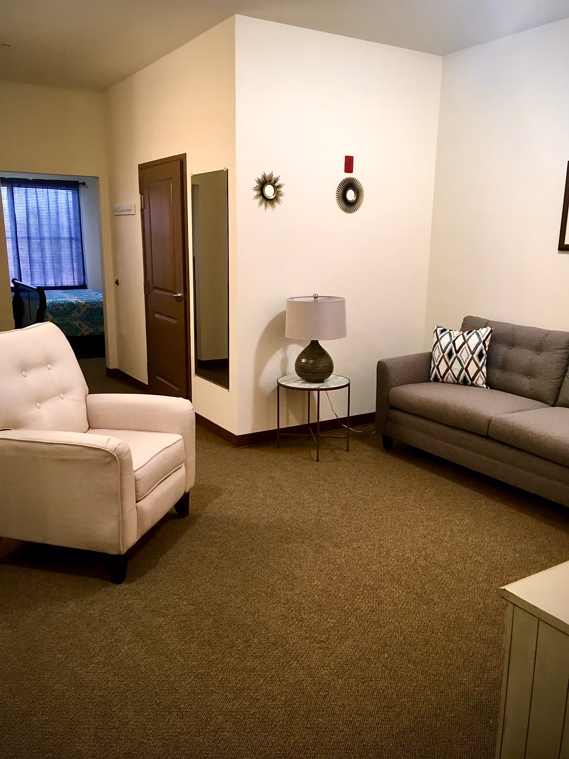 Well-lit living room with a beige armchair, gray tufted sofa, side table with a lamp, and a doorway leading to a bedroom.