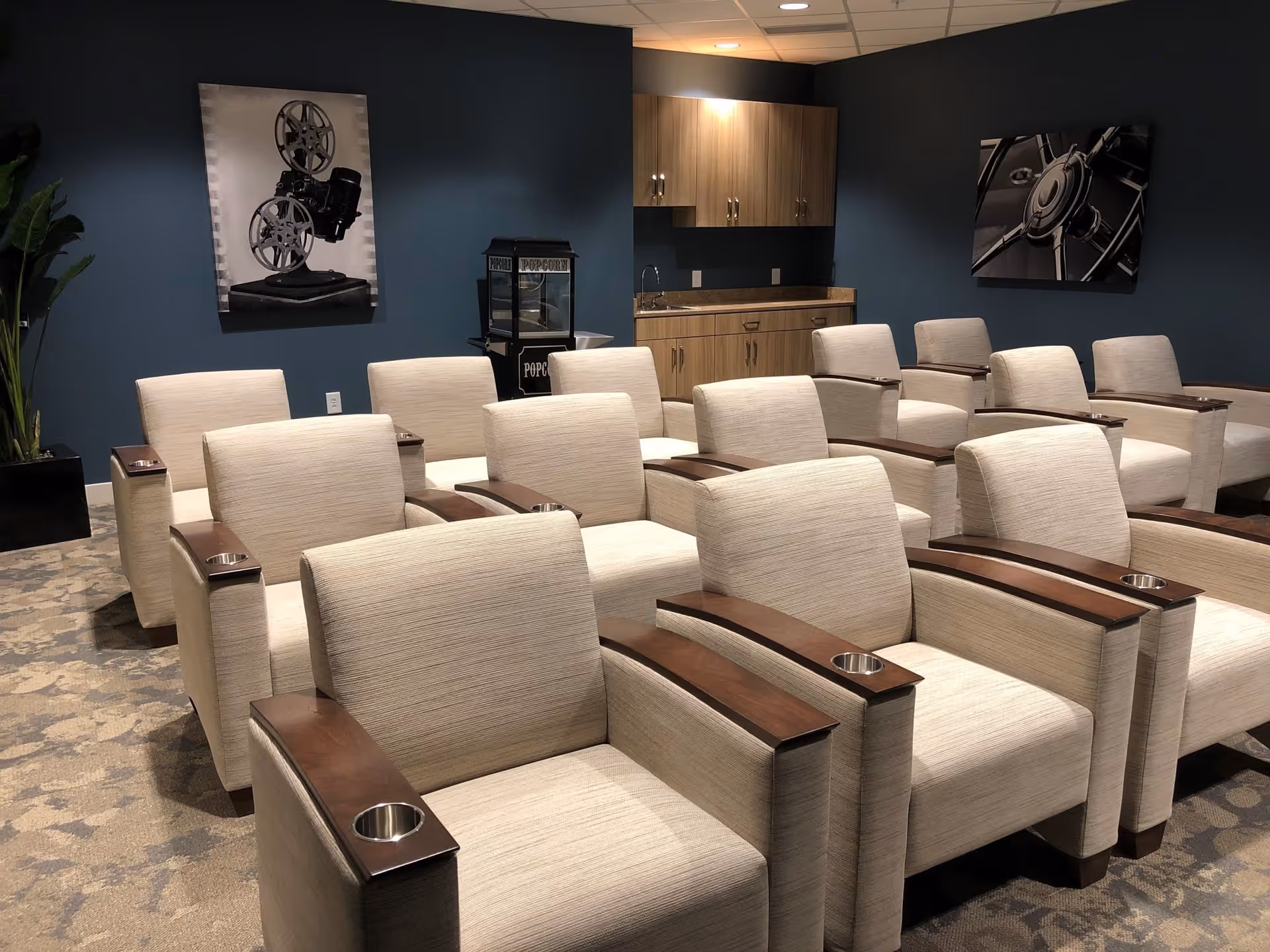 A small theater room with three rows of beige upholstered armchairs featuring wooden armrests with cup holders. The room has dark blue walls with two black and white framed pictures, a popcorn machine, and a small kitchenette with wooden cabinets and a sink in the back corner.