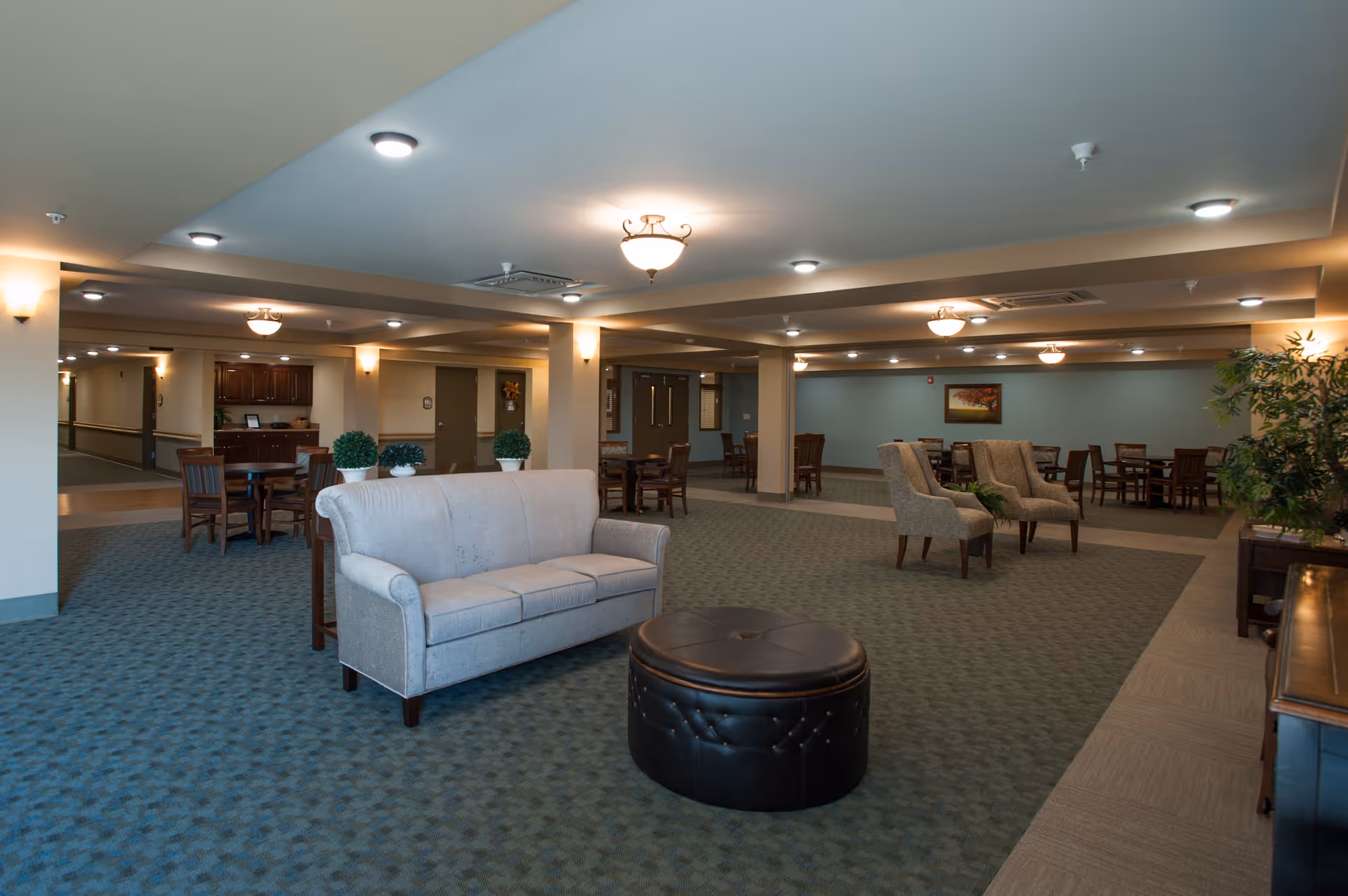 A spacious senior living facility common area with a light gray sofa and a round black ottoman in the foreground. Several wooden tables and chairs are arranged throughout the room, along with two upholstered armchairs. The room is well-lit with ceiling lights and wall sconces, and there are decorative plants and framed artwork on the walls.