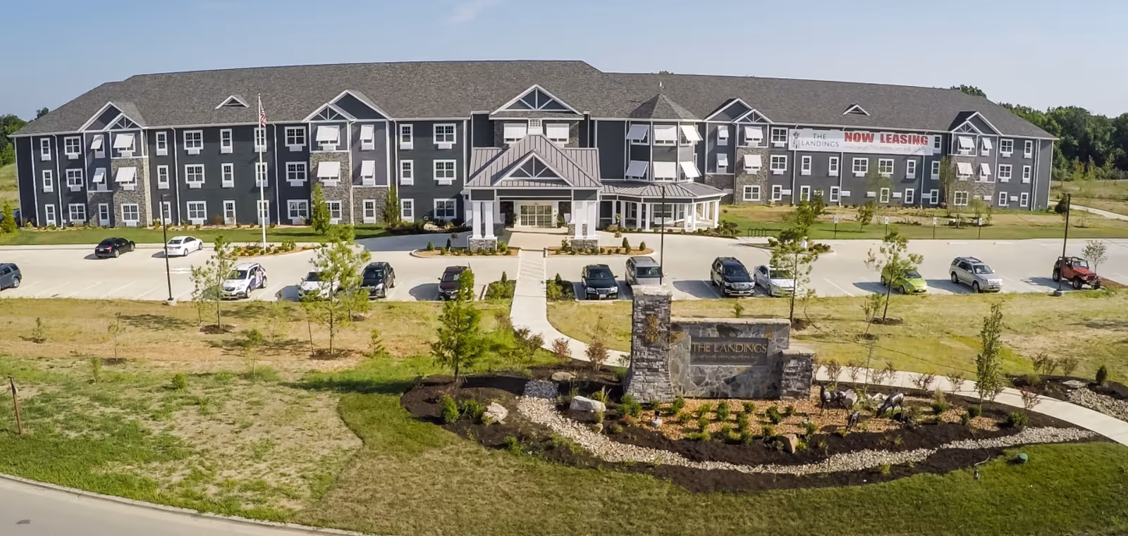 Front exterior view of The Landings at Reed Station Crossing, a large three-story building with multiple windows and white awnings. There is a covered entrance with pillars, a parking lot with several cars, a landscaped area with small trees and shrubs, and a stone sign displaying the facility's name. A banner on the building reads 'NOW LEASING'.