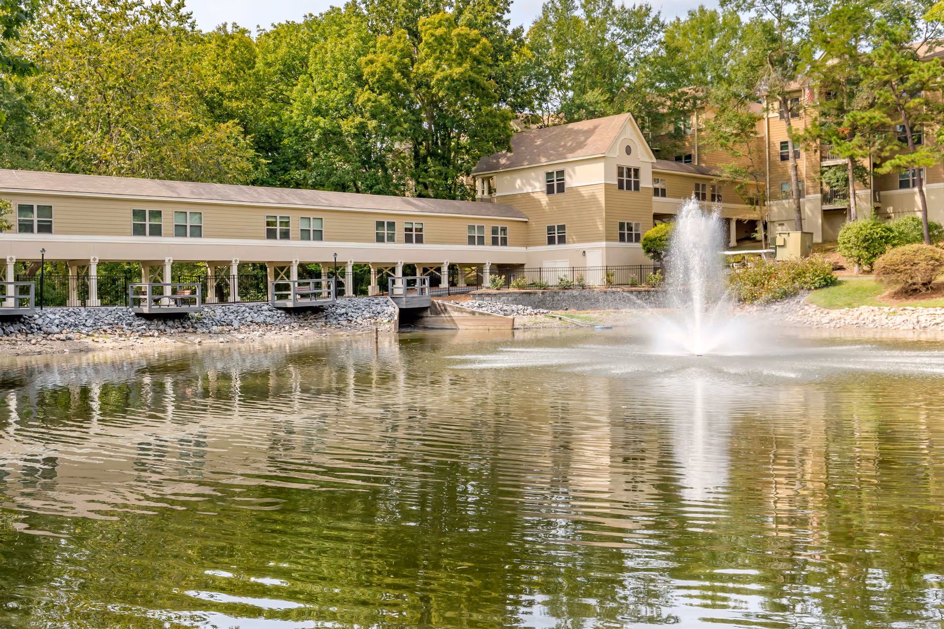 A serene outdoor scene at Brookdale Carriage Club Providence featuring a pond with a water fountain in the center, surrounded by trees and greenery. In the background, there is a beige multi-story building with a covered walkway extending over part of the pond.