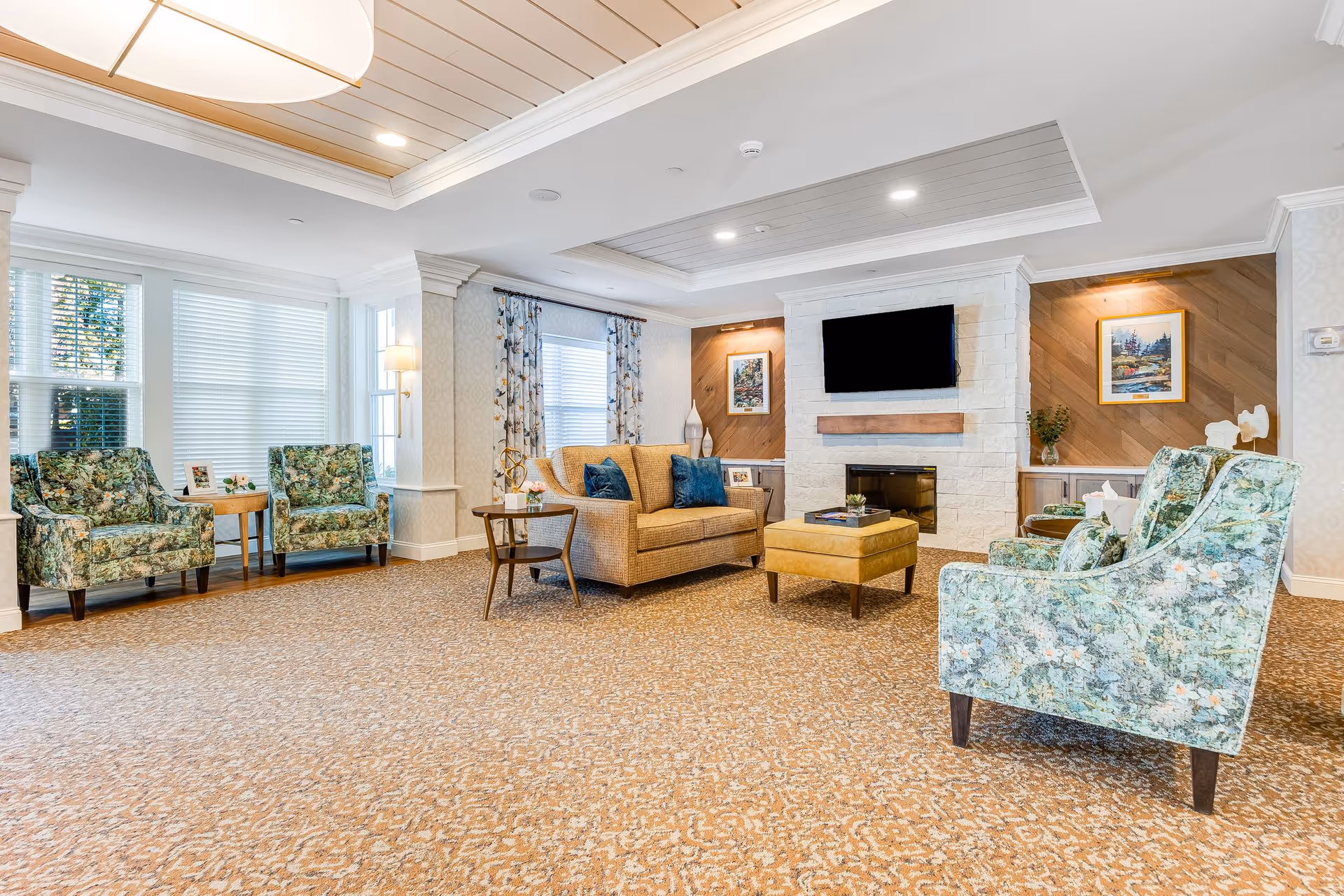 A bright and spacious living room area with floral patterned armchairs, a beige sofa with blue cushions, a yellow ottoman, and a fireplace with a mounted flat-screen TV above it. The room features large windows with blinds and floral curtains, decorative wall art, and a beige patterned carpet.