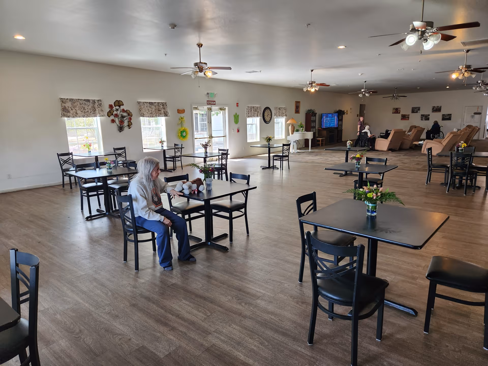 A spacious common area in a senior living facility with several black tables and chairs arranged on a wood floor. An elderly woman with gray hair is sitting at one of the tables holding a stuffed animal. In the background, there are reclining chairs, a television, and a piano. The room has multiple windows with floral curtains and ceiling fans with lights.