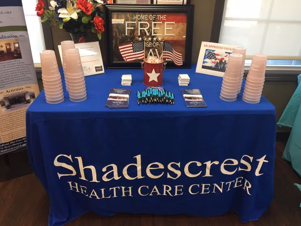 A promotional table covered with a blue tablecloth that reads 'Shadescrest Health Care Center'. The table holds stacks of pink plastic cups, brochures, pens, and a patriotic-themed decoration with American flags and a sign that says 'Home of the Free Because of the Brave'. There are informational posters and a window in the background.