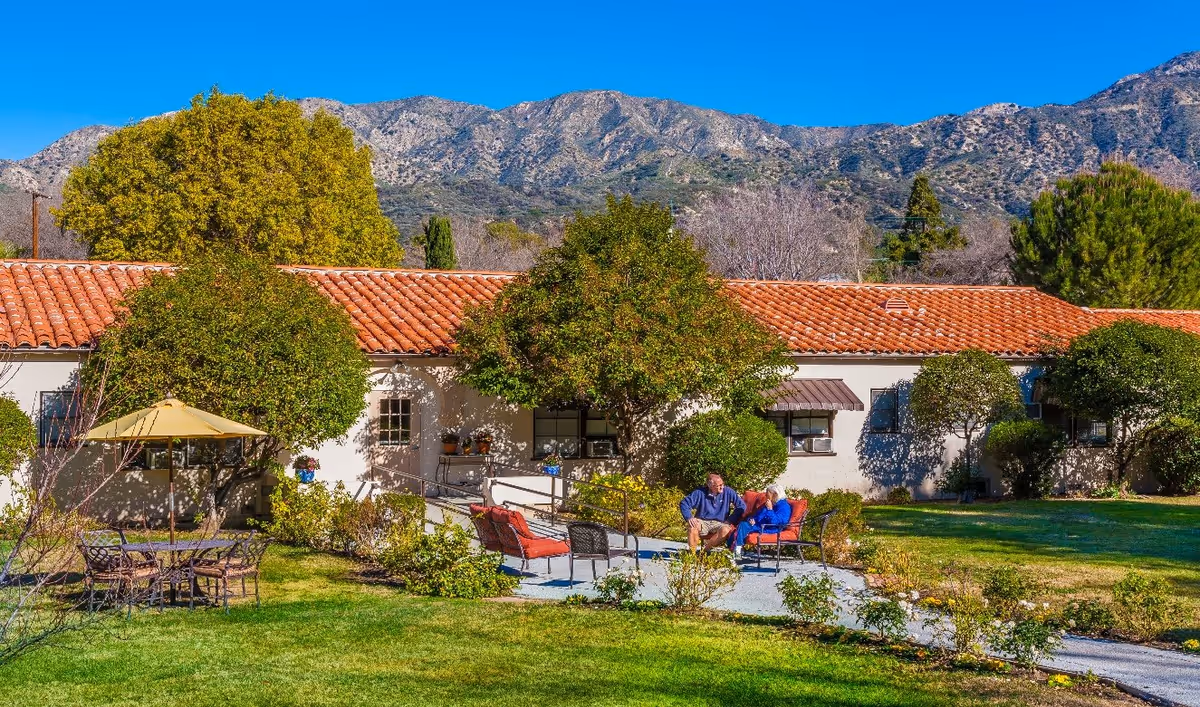 Outdoor patio area at Foothill Retirement with two people sitting and talking on cushioned chairs surrounded by greenery, trees, and a building with a red tile roof. Mountains are visible in the background under a clear blue sky.