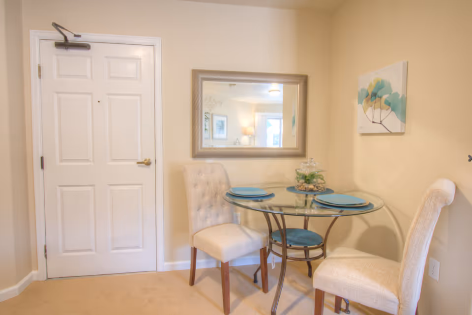 Small dining nook with a round glass table set for two, two upholstered chairs, a wall mirror, and an entry door.