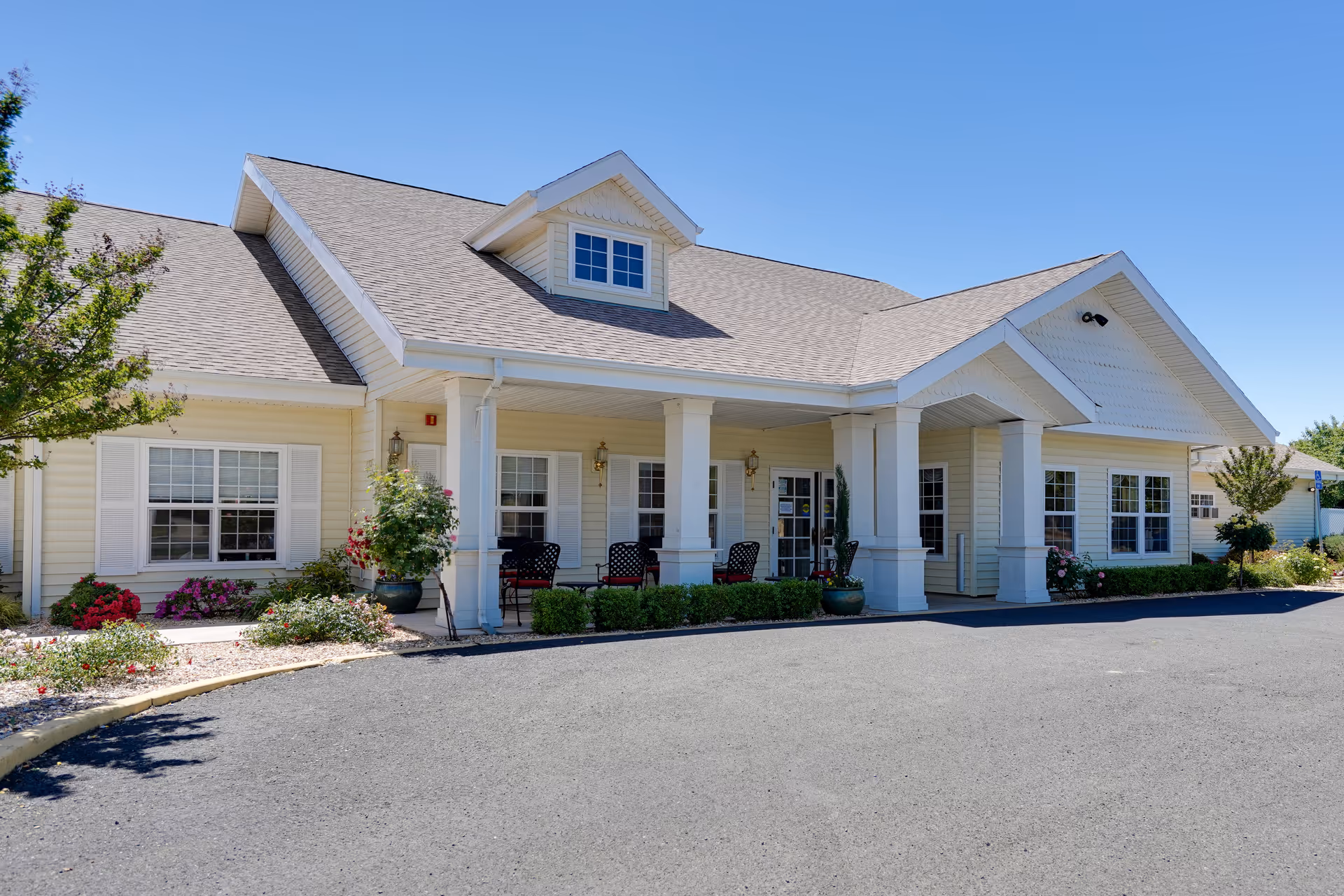 Exterior view of a single-story senior living community building with a covered entrance supported by white columns. The building has beige siding, white trim, and multiple windows with white shutters. There are potted plants and outdoor chairs near the entrance, and a clear blue sky above.