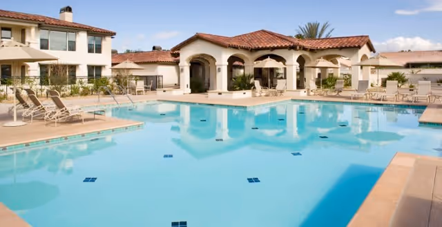 Outdoor swimming pool surrounded by lounge chairs and a building with arches and a red-tiled roof under a partly cloudy sky.