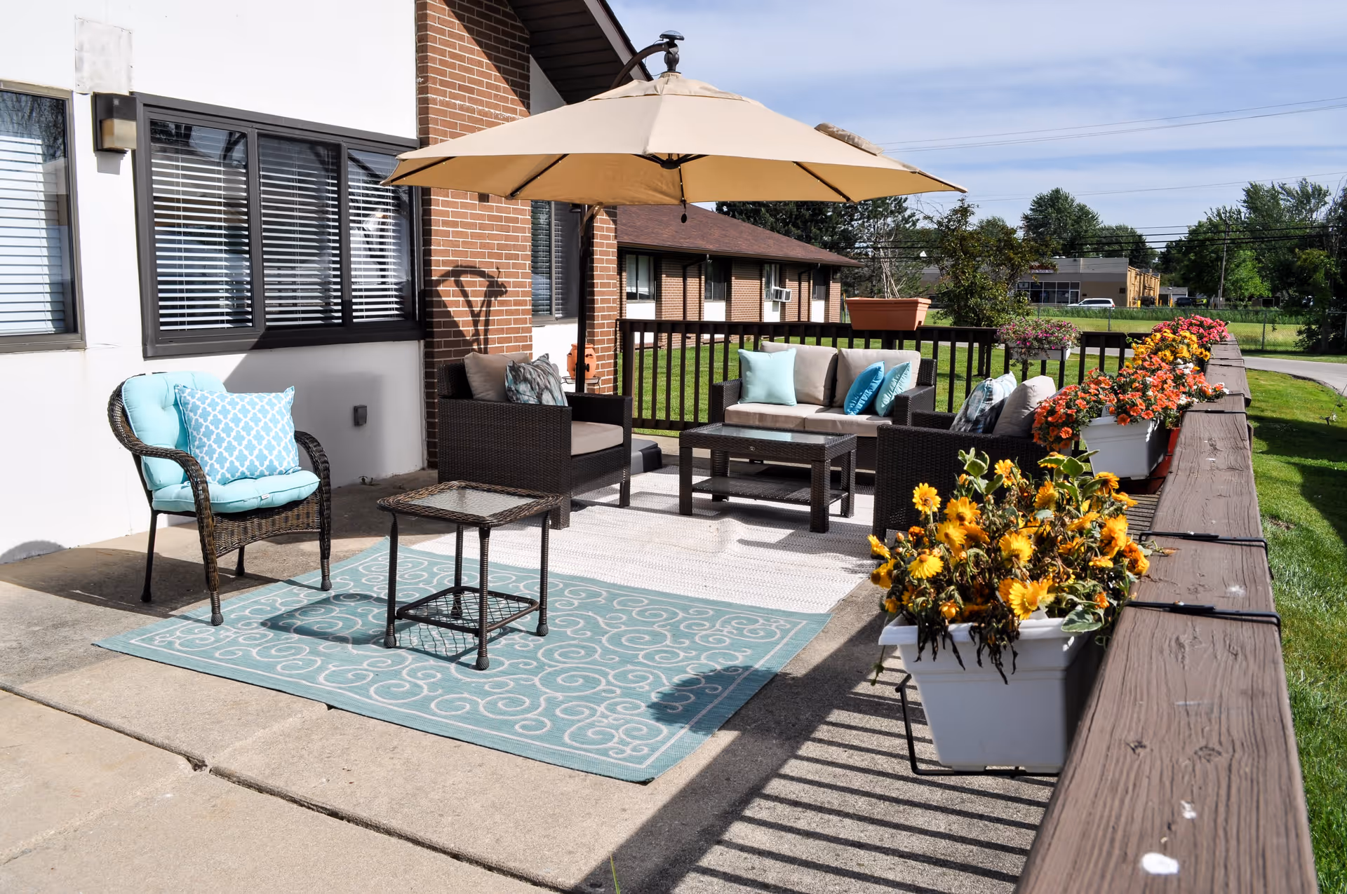 Outdoor patio area with wicker furniture including a loveseat, chairs with cushions, a small table, and a large beige umbrella. The patio is decorated with a blue patterned rug and several flower pots filled with colorful flowers along the wooden railing. The background shows a grassy area and neighboring buildings under a clear sky.