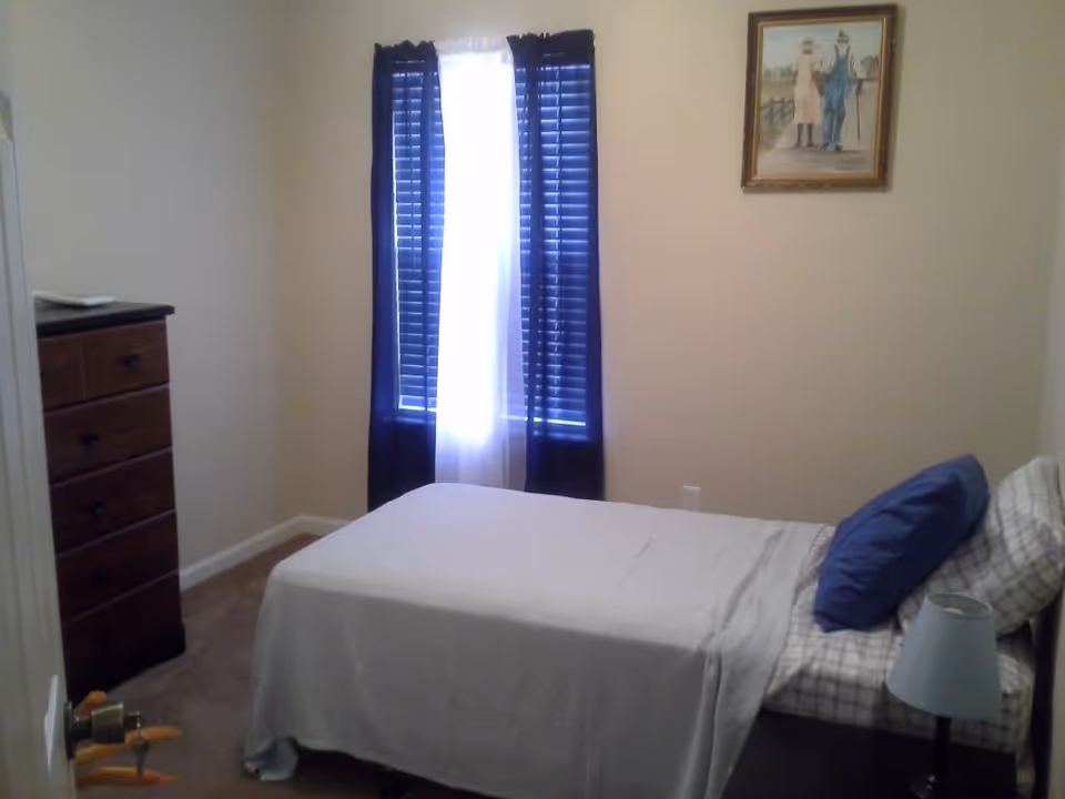 A small bedroom with a single bed covered with a white blanket and a blue pillow. There is a wooden dresser on the left side and a window with dark blue curtains letting in some light. A framed picture hangs on the wall above the bed, and a small bedside lamp is visible on the right side.