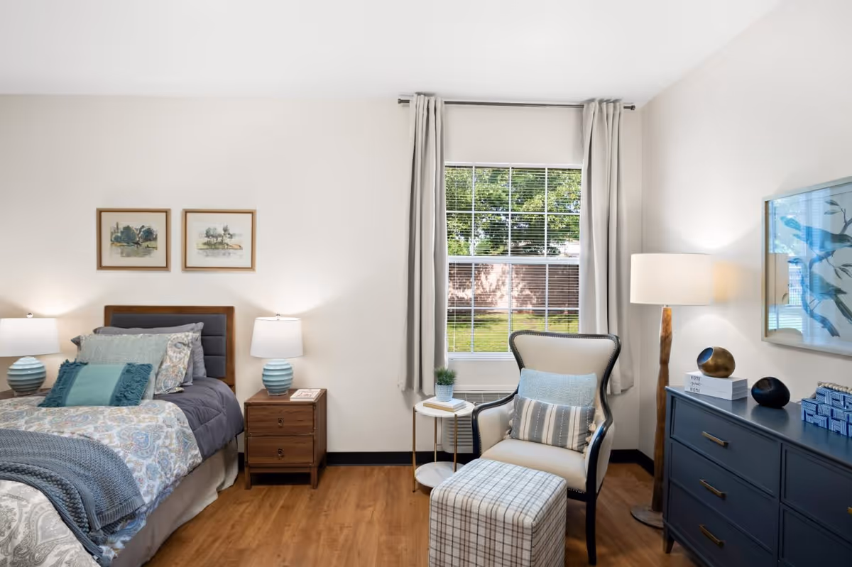 Bright, tastefully decorated bedroom with a bed, nightstand, armchair and ottoman beside a window overlooking a yard.