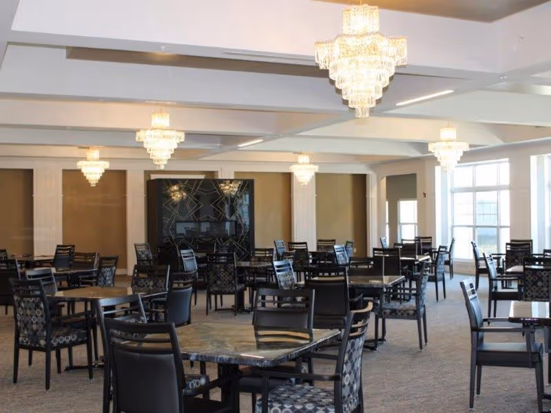 A spacious dining room with multiple tables and chairs arranged neatly. The room features large windows allowing natural light to fill the space, and elegant chandeliers hanging from the ceiling. The walls are decorated with columns and a large decorative panel.