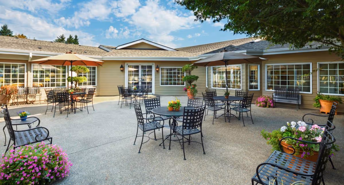 Outdoor patio area at Canterbury Inn Assisted Living Community with several black metal tables and chairs, some shaded by umbrellas. The patio is surrounded by beige building walls with large windows and decorated with potted plants and flowers. A tree provides additional shade on the right side.