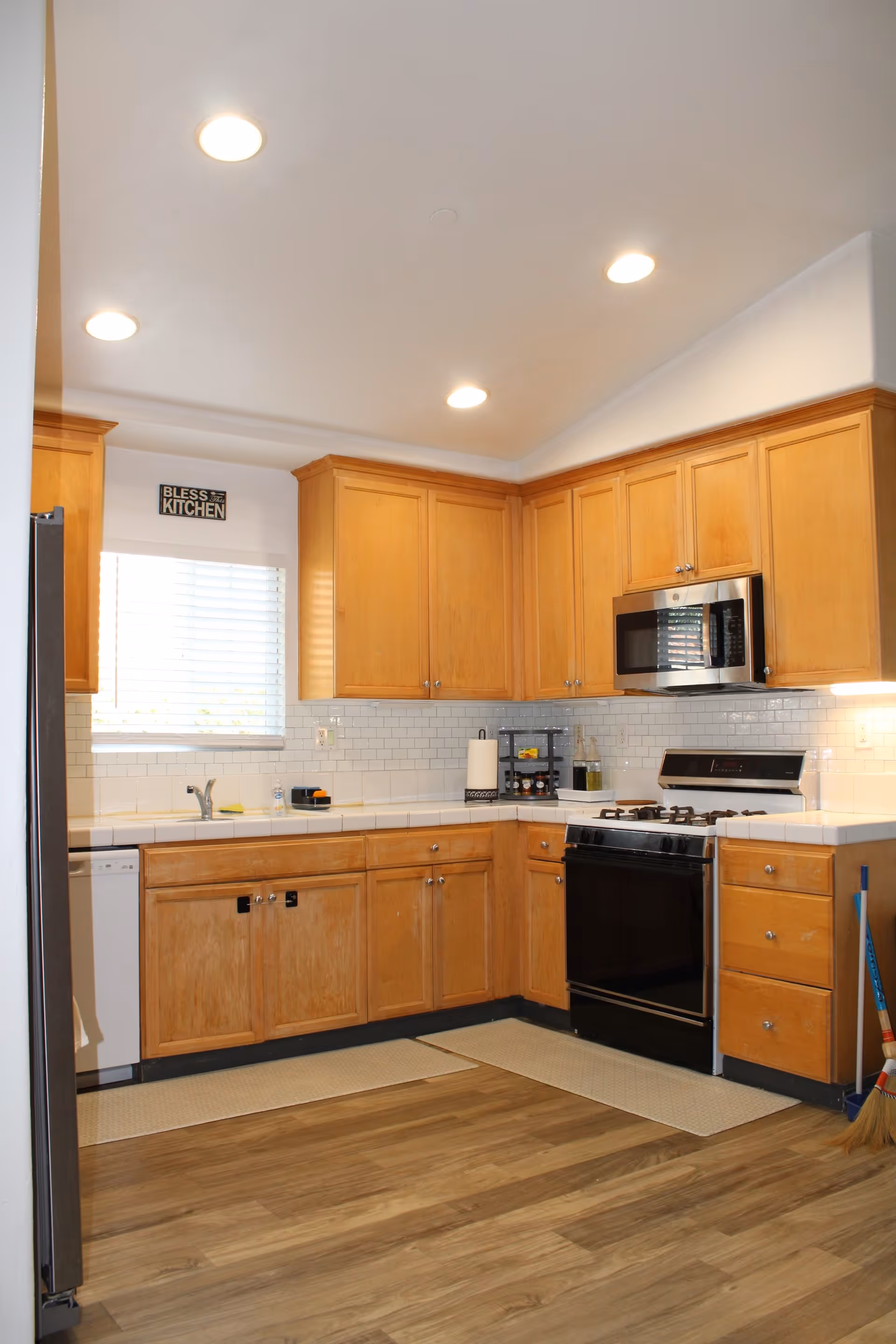 A clean kitchen with wooden cabinets, white tiled backsplash, a black stove with oven, a microwave above it, a dishwasher, and a window with blinds. The floor is wooden with two beige rugs, and there is a broom and dustpan in the corner. A small sign above the window reads 'Bless this Kitchen'.