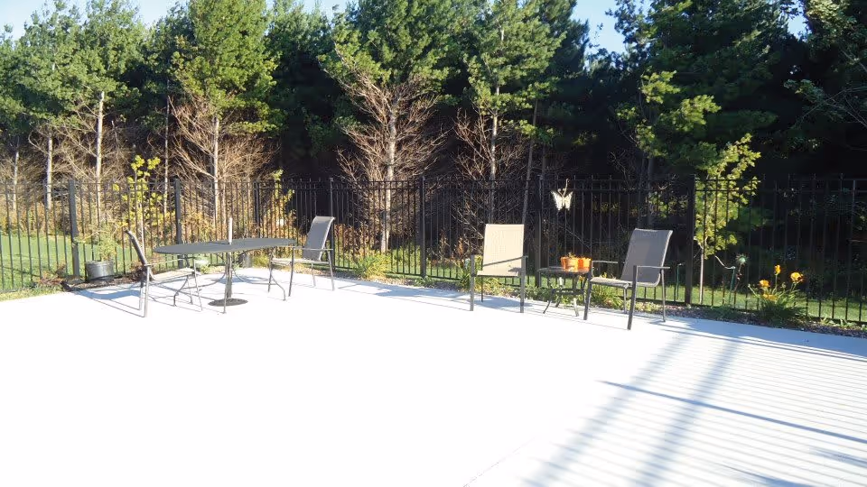Outdoor patio area with a concrete floor, featuring two sets of metal tables and chairs. The patio is enclosed by a black metal fence, with trees and greenery visible beyond the fence under a clear blue sky.