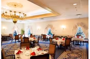 Formal dining room with tables set with white tablecloths and red napkins, chandeliers, and draped windows.