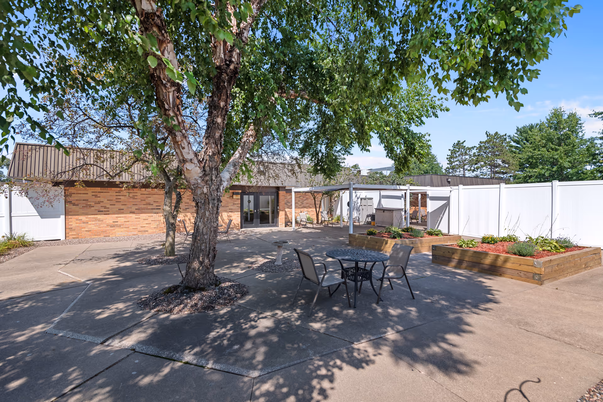 Outdoor patio area with a large tree in the center, surrounded by concrete pavement. There are two raised garden beds with plants and mulch, a small round metal table with two chairs, and a building with brick walls and a brown roof in the background. The area is enclosed by a white fence and there is a clear blue sky above.