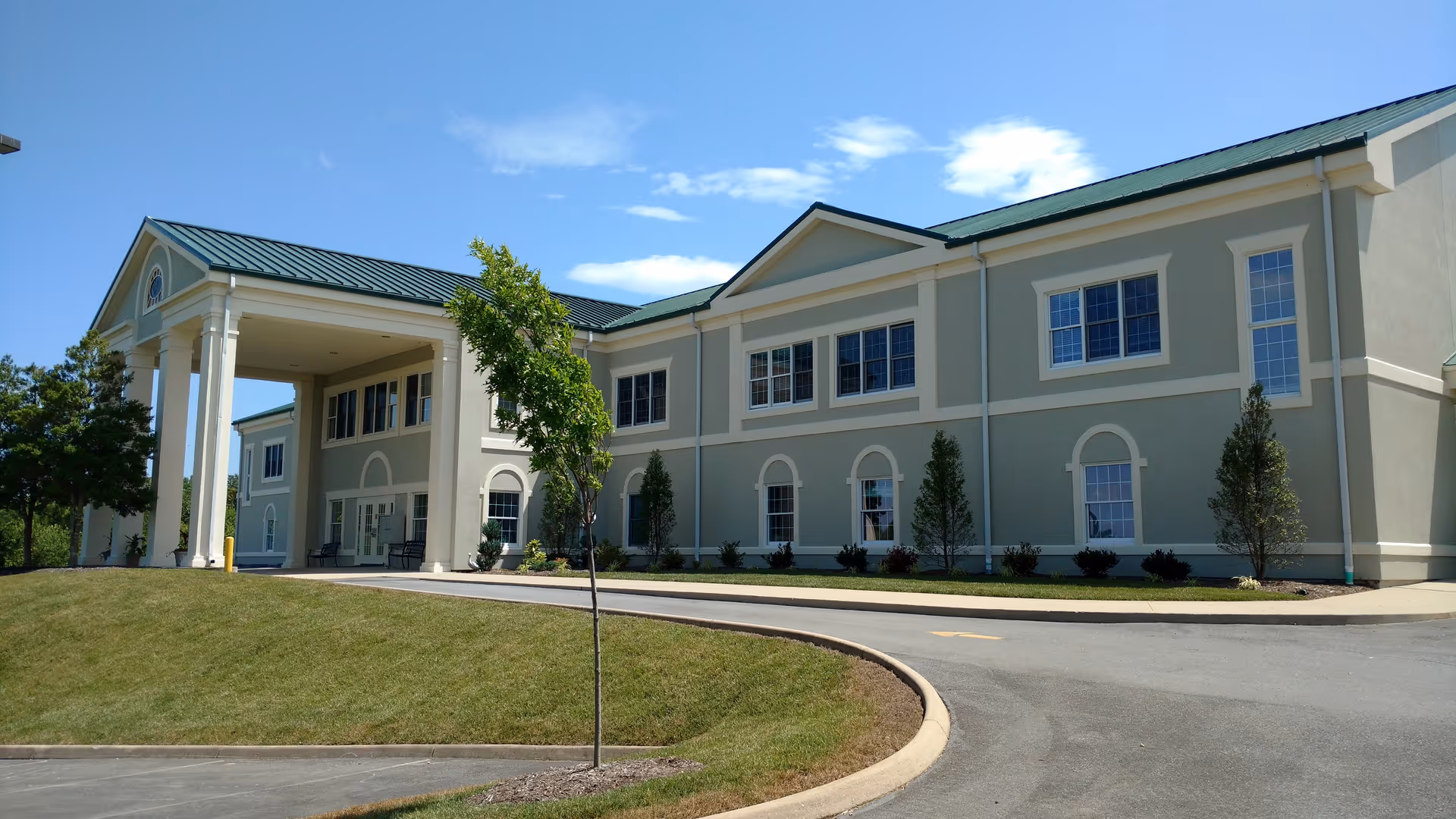Exterior view of a two-story retirement and assisted living facility building with a covered entrance supported by columns, green metal roof, multiple windows, and a curved driveway with landscaped grass and small trees.
