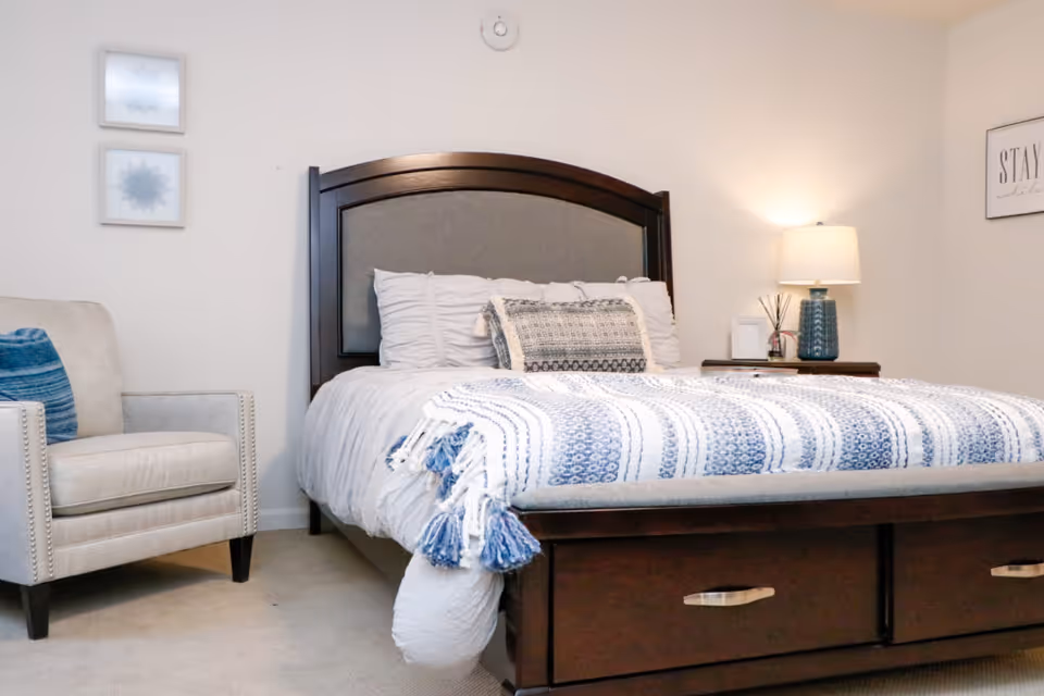 Well-lit bedroom with a bed featuring blue-and-white bedding, a dark wood headboard and under-bed drawers, a beige armchair, and a nightstand with a lamp.