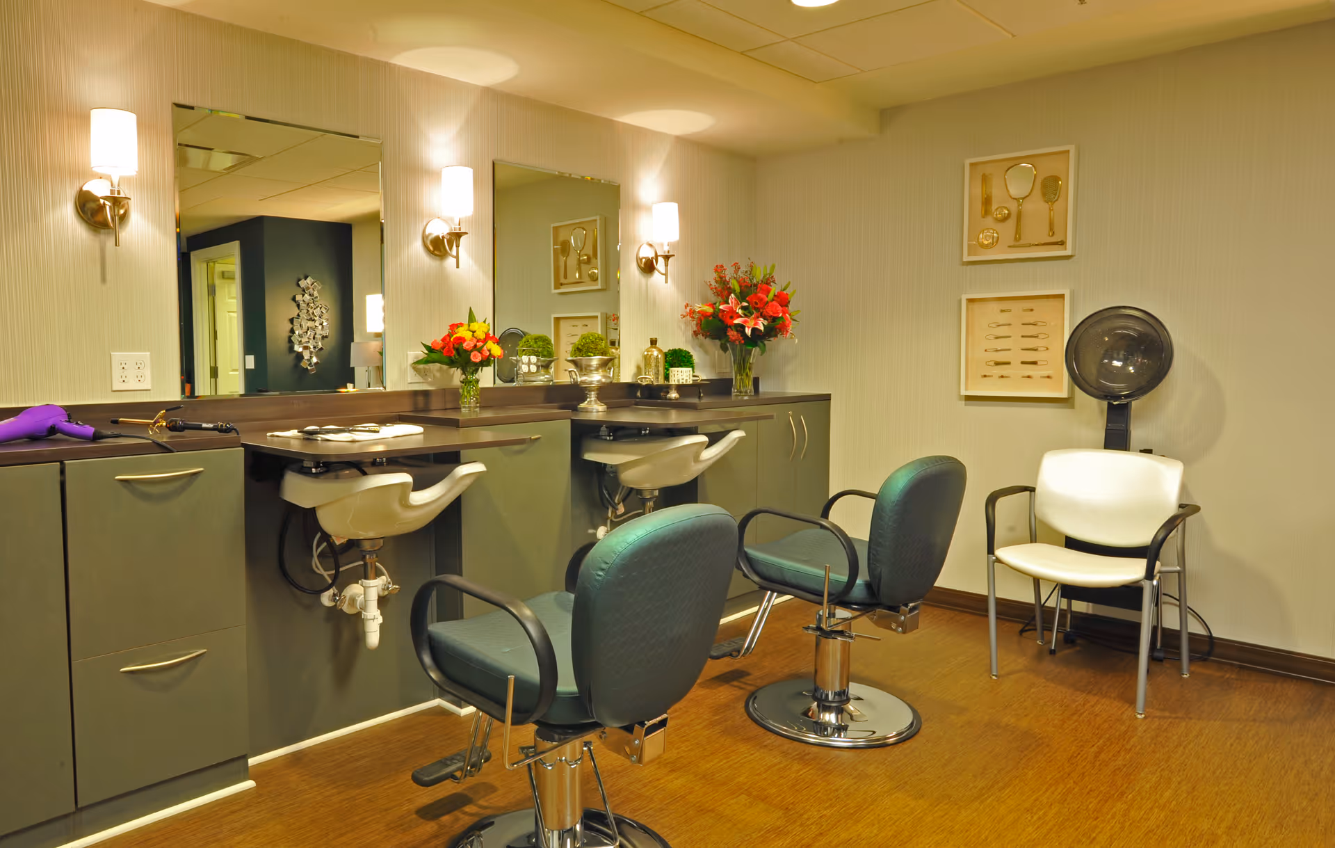 Interior of a salon room with two styling chairs in front of a counter with mirrors and wall-mounted lights. There are flowers on the counter, a hair dryer, and a white chair next to a hair drying hood. The walls have framed pictures of vintage grooming tools.