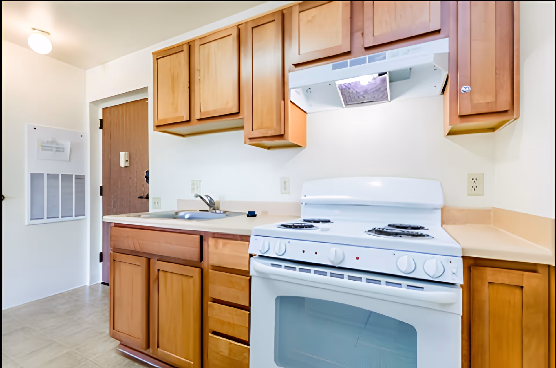 A small kitchen area with wooden cabinets, a white electric stove with four burners, a range hood, a sink with a faucet, and a wooden door in the background. The walls are white and the floor has light-colored tiles.