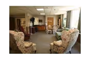 Cozy communal living room with floral armchairs, a sofa, a small dining table, TV, and large windows.