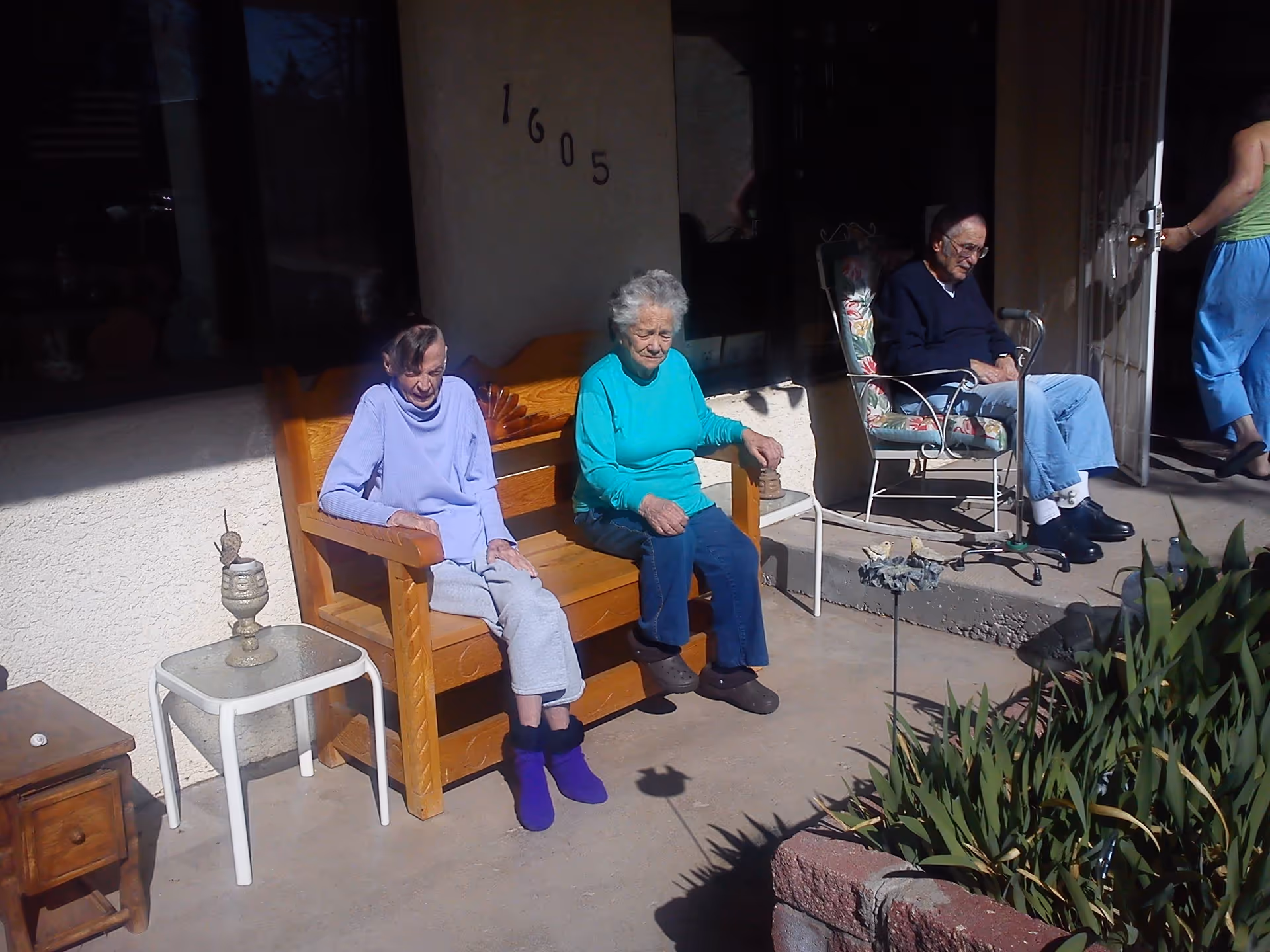 Two elderly women sitting on a wooden bench outside a building with the number 1605 on the wall. Another elderly man is sitting on a floral cushioned metal chair nearby, and a person in blue pants and a green top is walking through a door in the background. There are plants and garden decorations in the foreground.