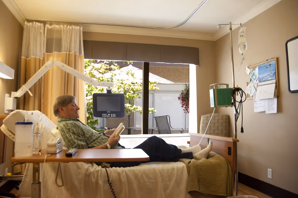 An older man reclines in a medical-style bed in a bright patient room, holding a remote with IV equipment nearby and a window looking out to a patio.