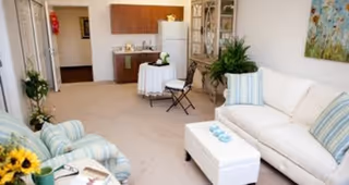 A bright and cozy living room area featuring a white sofa with striped blue and white pillows, a matching armchair, a white ottoman with decorative items on top, a small round table with two chairs near a kitchenette area with wooden cabinets and a white refrigerator, and a large plant in the corner. The room has light-colored carpet and walls with artwork hanging above the sofa.