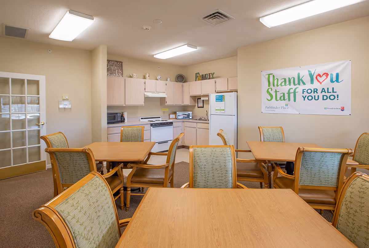Communal dining area with wooden tables and chairs and a small kitchen in the background with a 'Thank You Staff' banner on the wall.