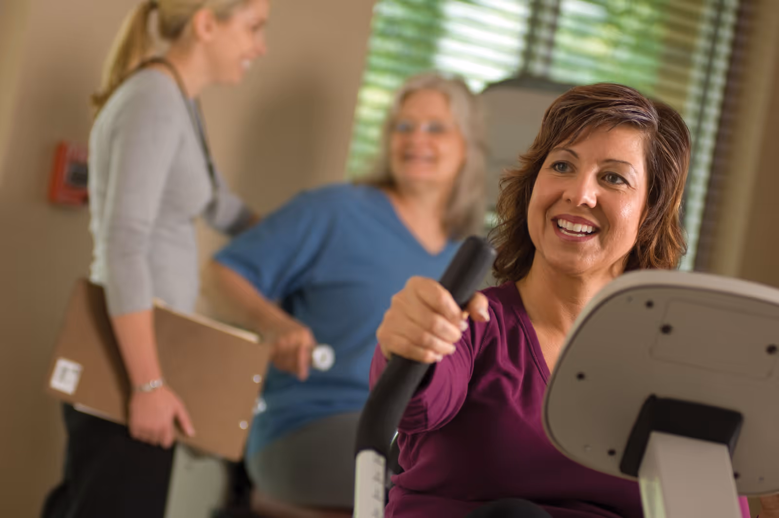 A woman in a purple top is using an exercise machine indoors, smiling. In the background, a woman in a blue shirt is seated and another woman holding a clipboard is standing, all appearing engaged in a fitness or rehabilitation activity.