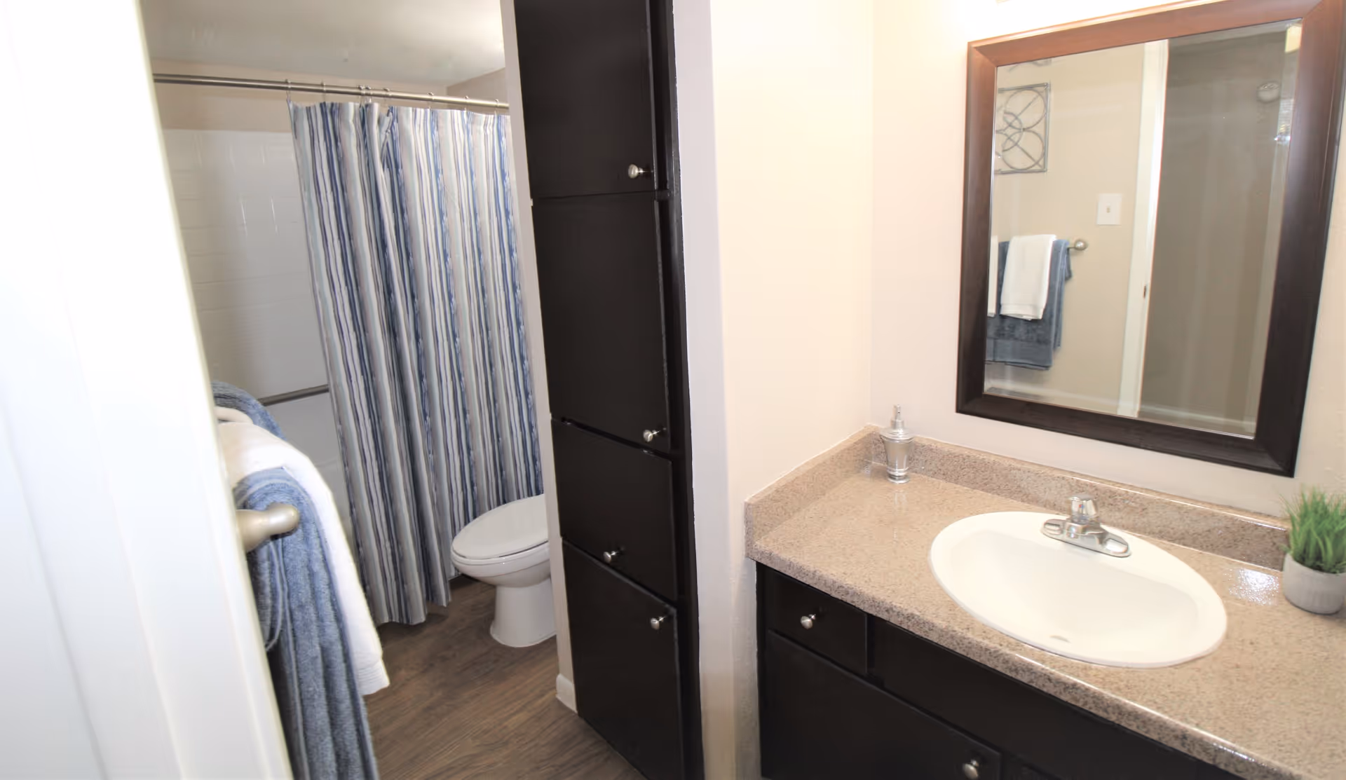 Bathroom with a sink and countertop on the right, a mirror above the sink, dark wood cabinets, a toilet partially visible behind a wall, and a shower with a blue and white striped curtain. Towels are hanging on a rack near the shower.
