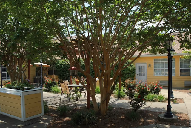 Outdoor courtyard area with trees, flowering bushes, a table with chairs, a bird feeder, and a yellow building with windows and a door in the background.