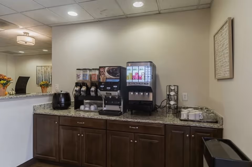 A beverage station with multiple drink dispensers including coffee and juice machines on a granite countertop with dark wooden cabinets below. There are cups and a cup holder on the right side, and a small black appliance on the left. The background shows a light-colored wall with a framed picture and ceiling lights.