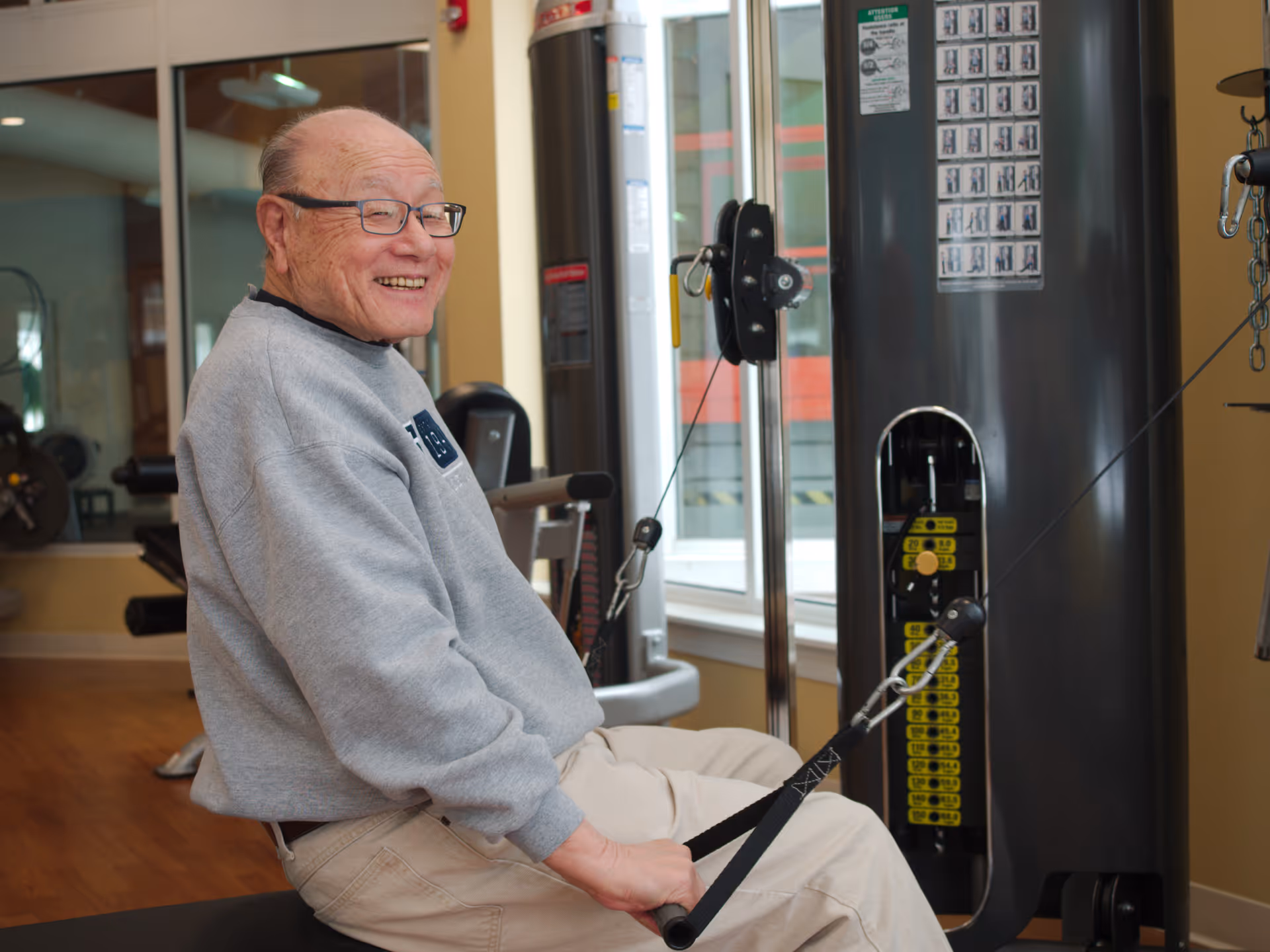 An elderly man wearing glasses and a gray sweatshirt is smiling while using a seated cable row exercise machine in a fitness room with large windows and gym equipment in the background.