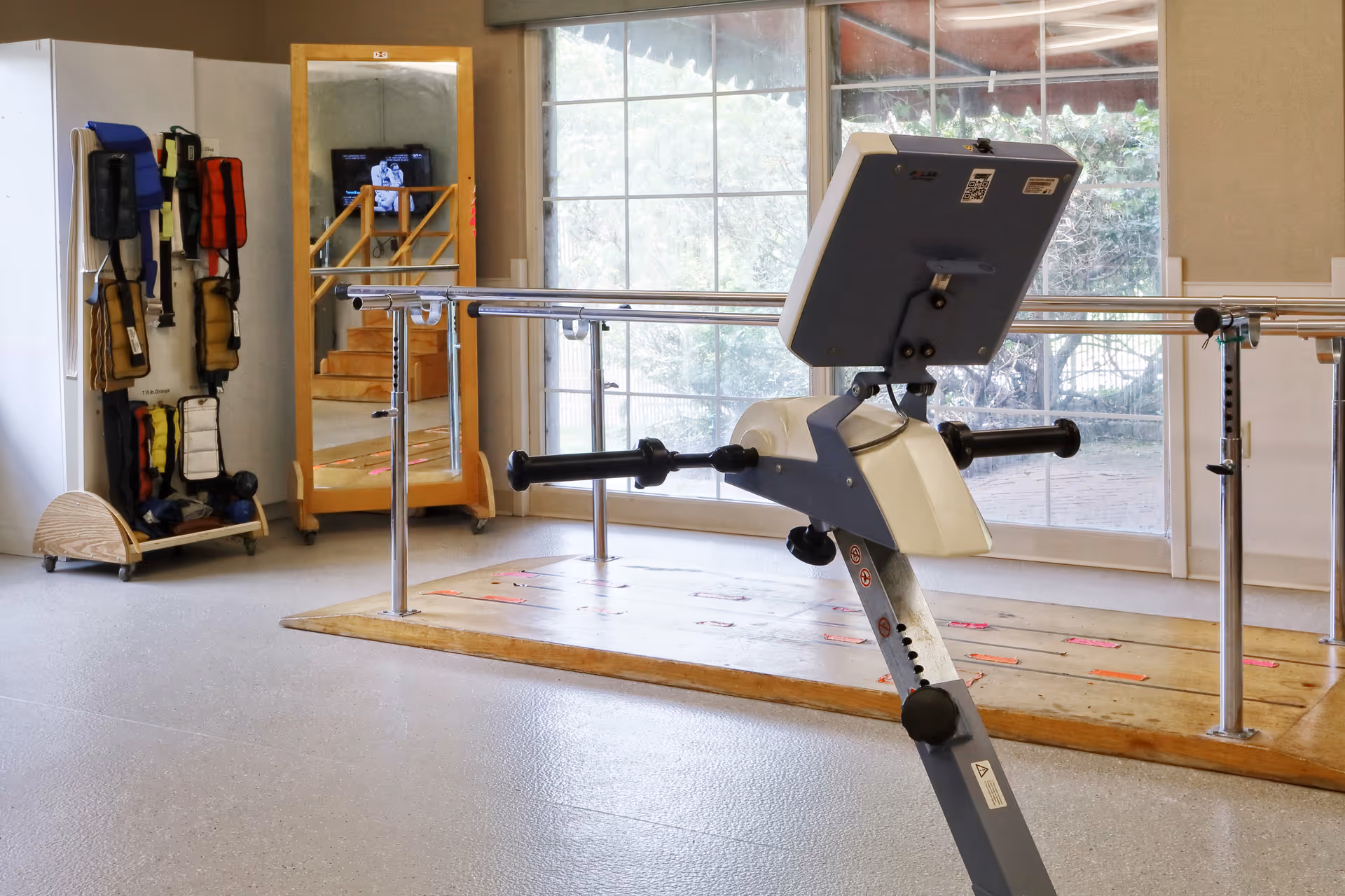 A physical therapy or rehabilitation room with parallel bars for walking exercises, a stationary exercise bike, a large mirror, and various weighted exercise belts hanging on a rack. Large windows provide natural light and a view of outdoor greenery.