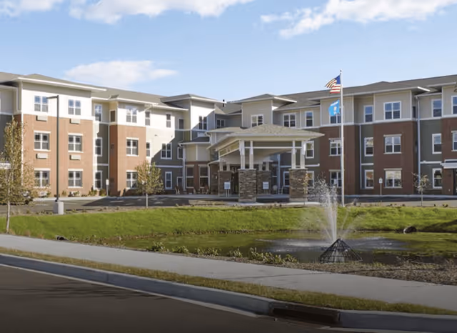 Exterior view of a multi-story senior living facility building with a covered entrance, landscaped lawn, small pond with a water fountain, and two flagpoles displaying the American flag and another flag under a partly cloudy sky.