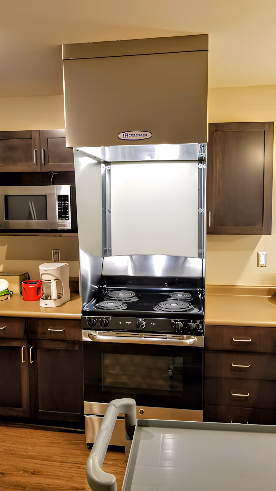A kitchen area with a stainless steel stove and oven, a large range hood above it, wooden cabinets on either side, a microwave on the left countertop, a coffee maker, and a red container. The floor is wooden, and a part of a gray cart is visible in the foreground.