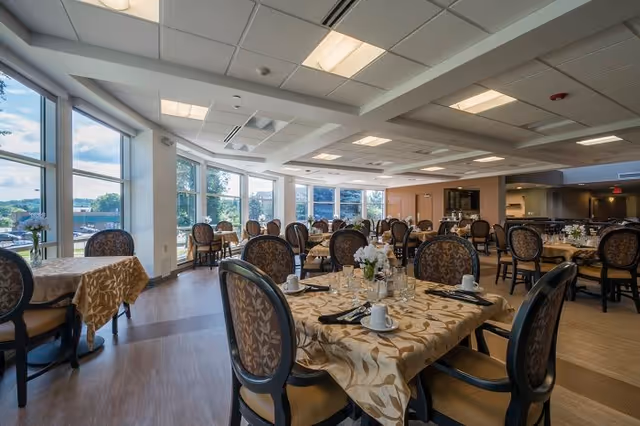 A spacious dining room with multiple round tables covered with beige patterned tablecloths. Each table is set with white cups, glasses, silverware, and a small floral centerpiece. The room has large windows allowing natural light to fill the space, and elegant cushioned chairs with patterned backs surround the tables. The ceiling has recessed lighting and a modern design.