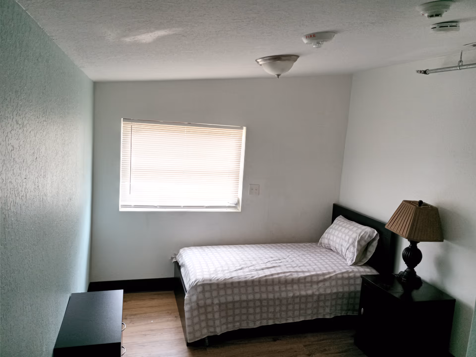 A small bedroom with a single bed covered in a checkered bedspread, a pillow, a bedside table with a lamp, a window with closed blinds, and a small black dresser against a light green textured wall.