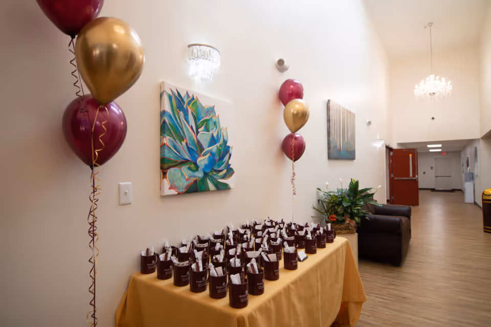 A hallway in Parkland Manor decorated with maroon and gold balloons. A table covered with a yellow tablecloth holds multiple maroon mugs filled with wrapped items. The walls are adorned with colorful paintings and there are plants and a brown armchair along the hallway. The floor is wooden and the ceiling has chandeliers.